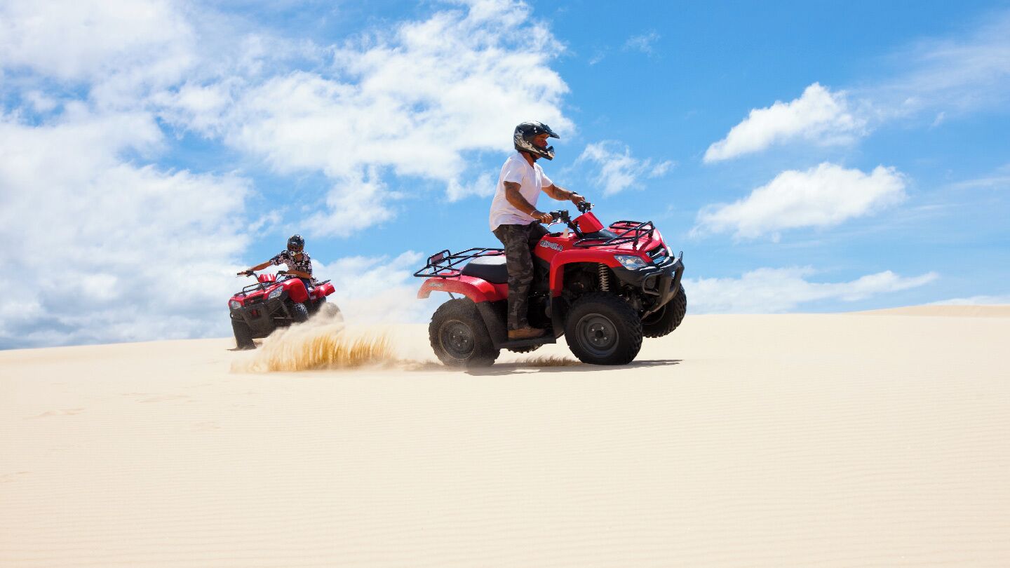 Zwei Personen fahren mit Quads eine Sanddüne bei blauem Himmel.