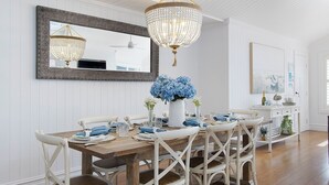 A dining area with a wooden table, white upholstered chairs, a chandelier, and a mirror on the wall.