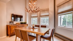 A dining area with a wooden table and chairs, a chandelier, a TV, and windows with a view of trees.