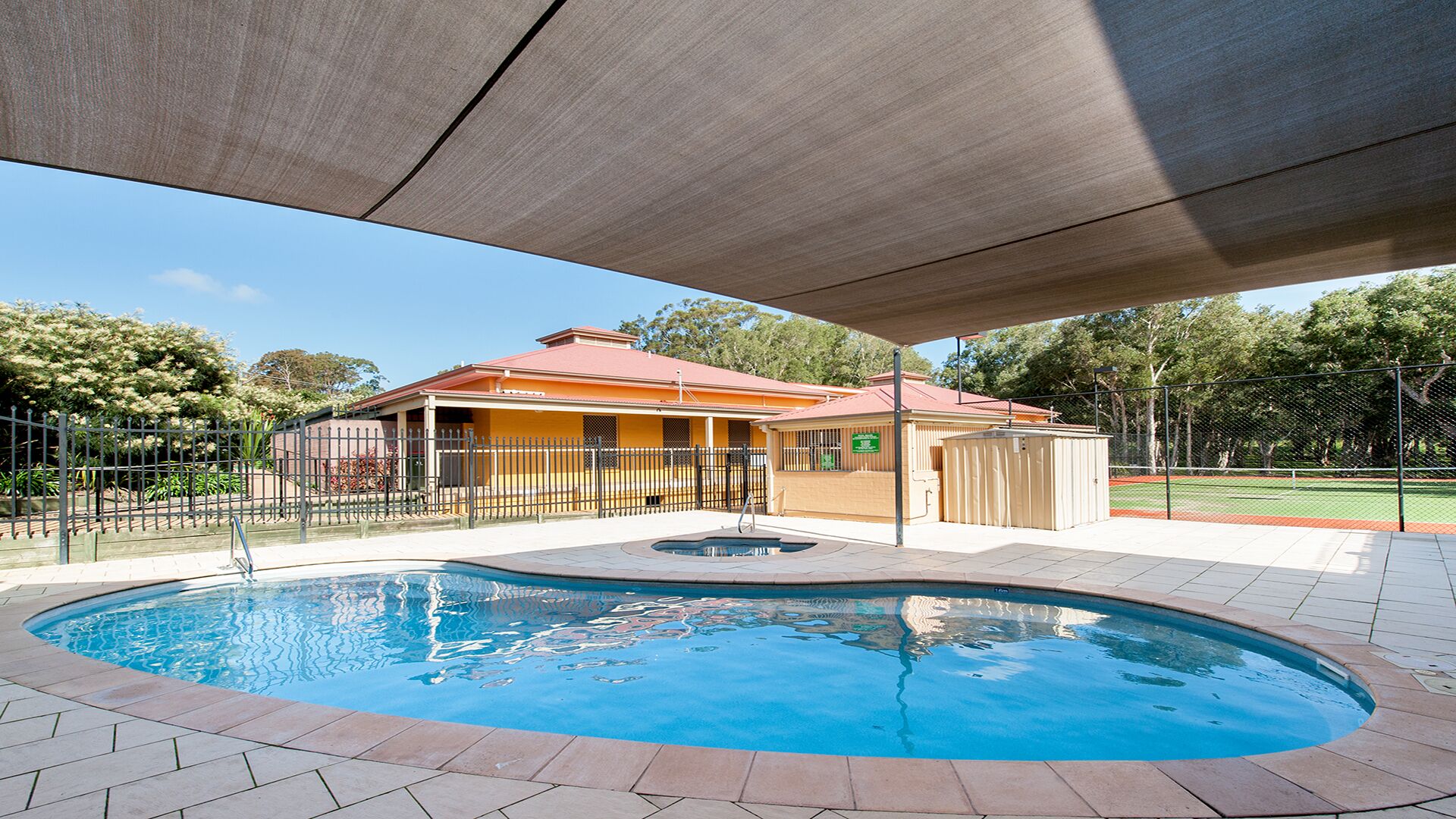 En swimmingpool med fliseterrasse, et overdækket område med tag, og en indhegnet have med træer samt en bygning i baggrunden.