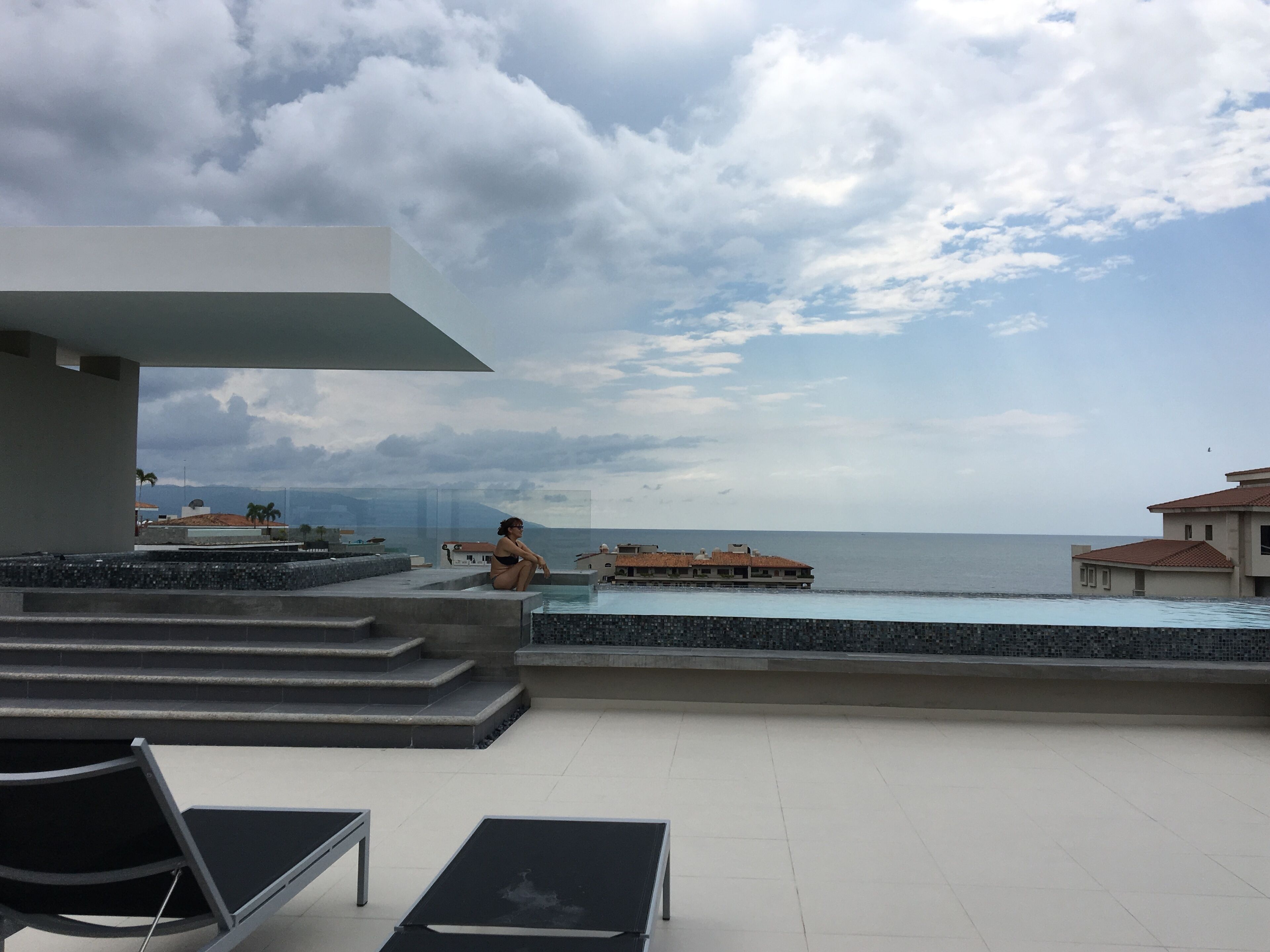 A rooftop pool area with lounge chairs, a view of the sea, and a building with a tiled roof.