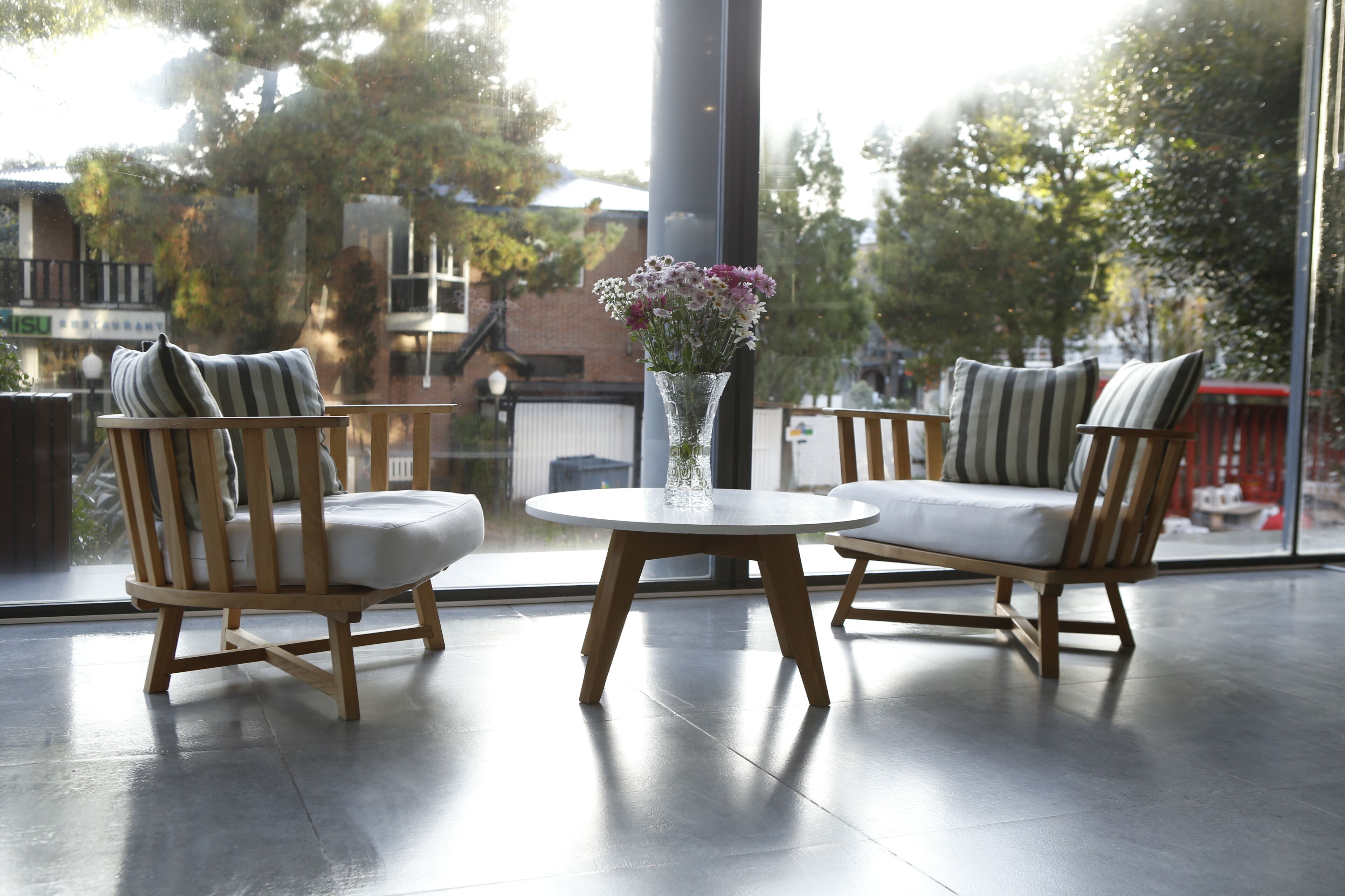 Un espace de détente intérieur moderne, avec deux chaises en bois et une petite table, un vase avec des fleurs et une grande fenêtre donnant sur des arbres et des bâtiments.