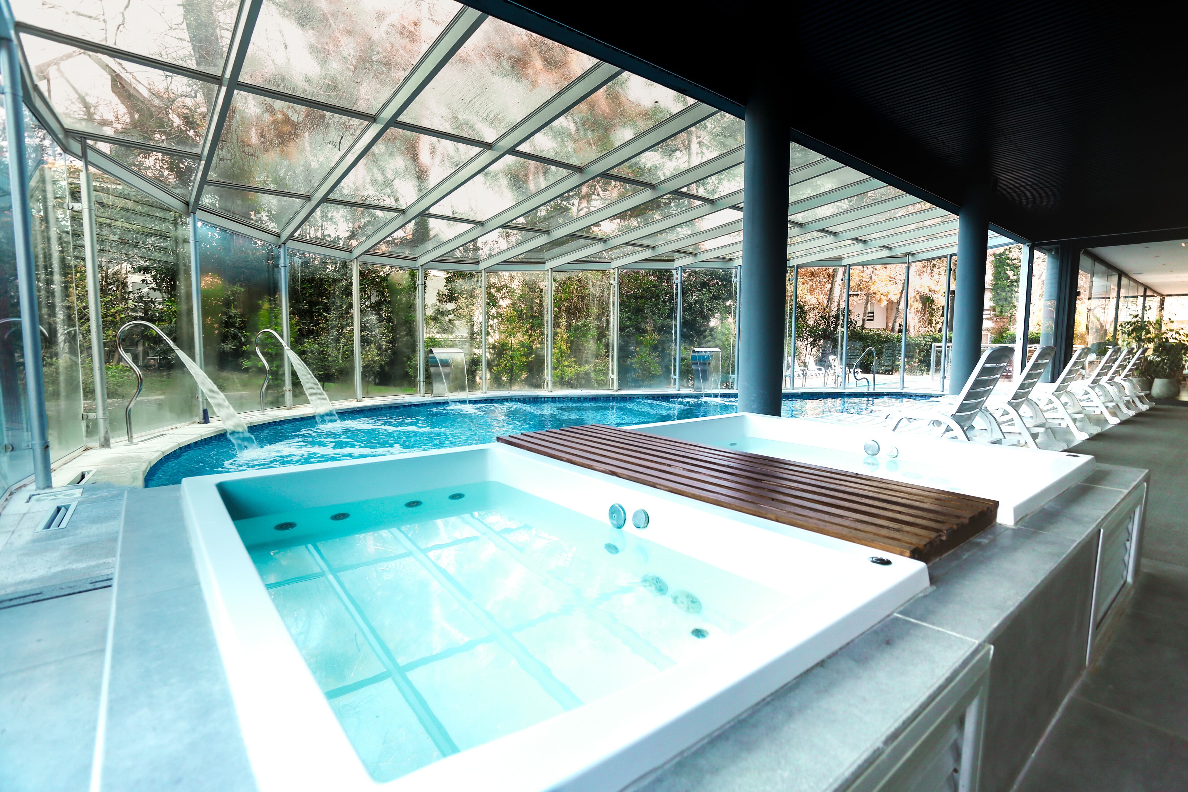 A modern indoor swimming pool with a glass ceiling, a hot tub, and lounge chairs.