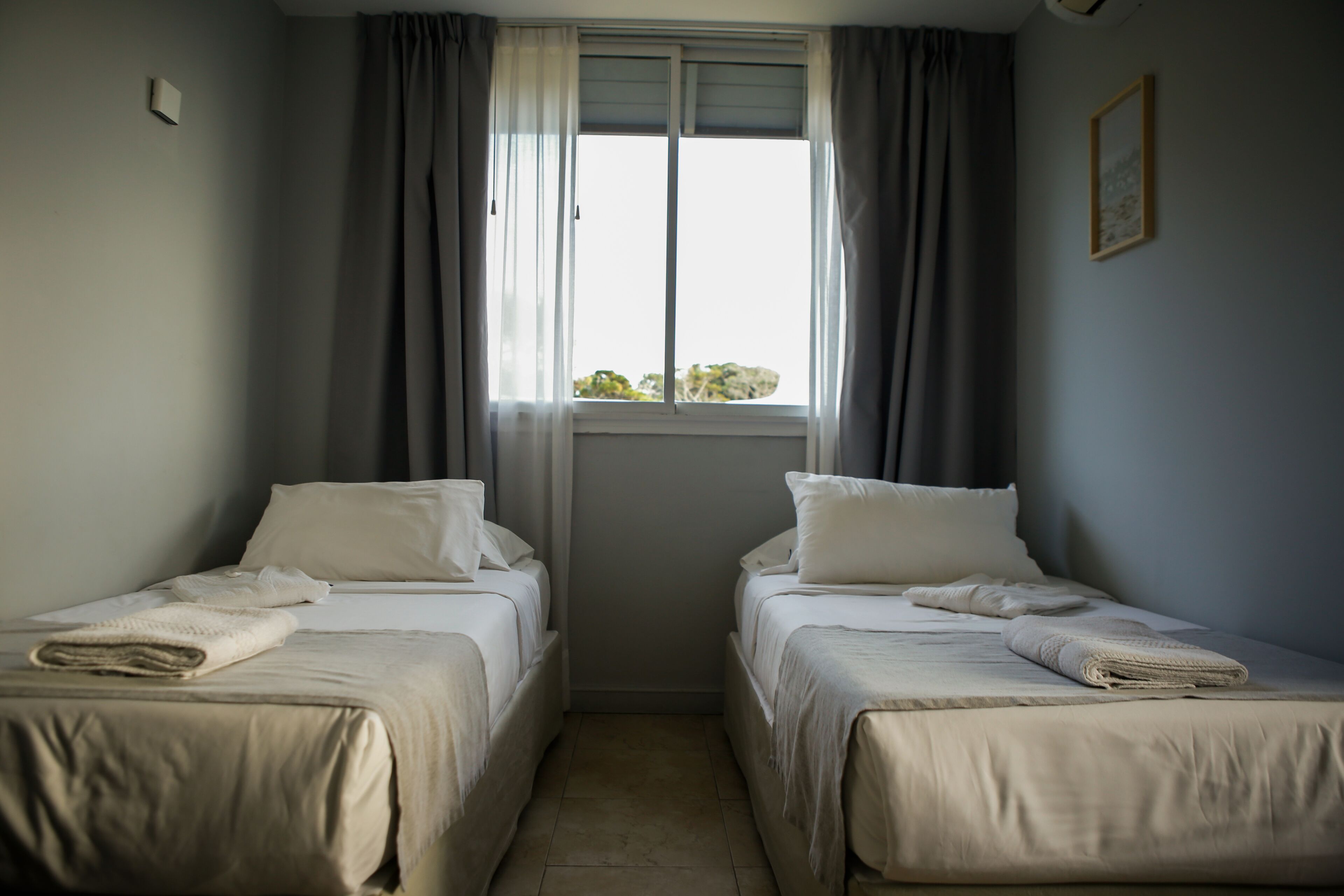 A room with two beds, a window with curtains, a small table, and a wall-mounted light.