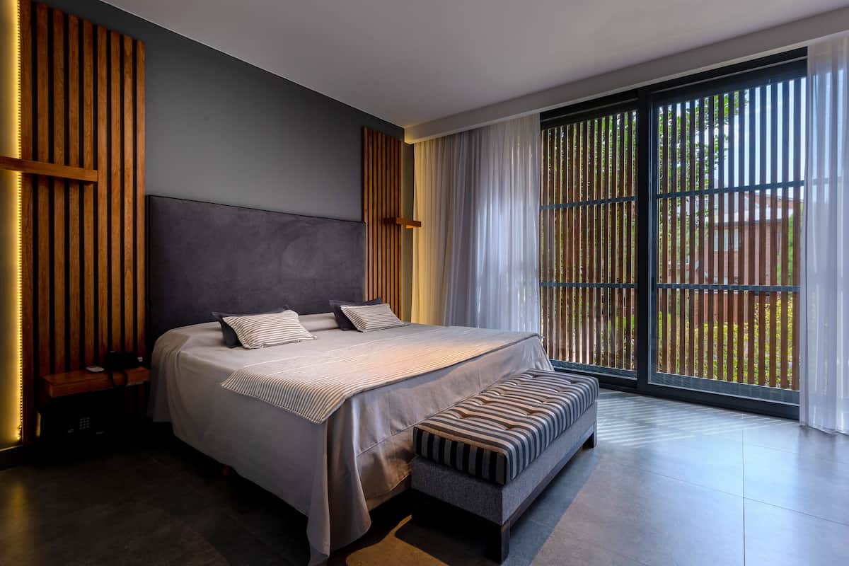 A modern bedroom with a large bed, a bench, and a view of greenery through large windows.