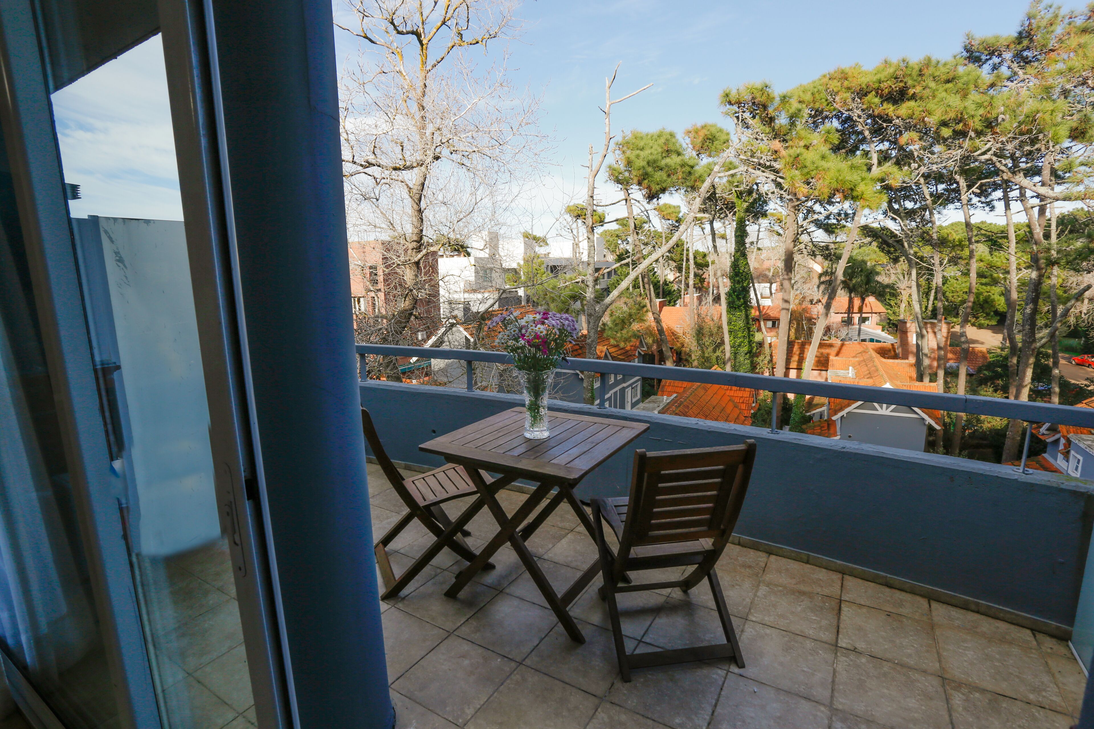 A balcony with a table and chairs, overlooking a residential area with trees and buildings.
