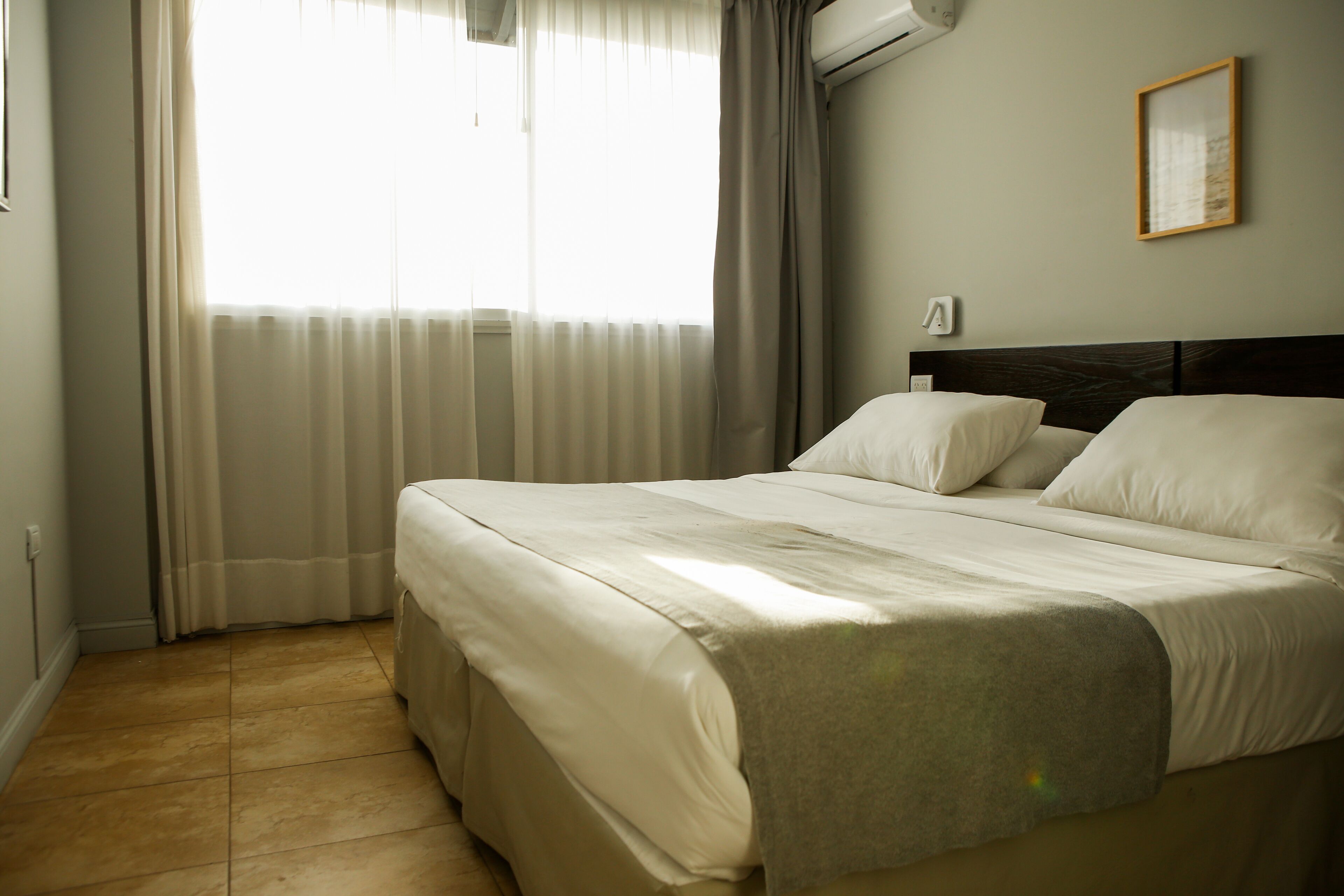 Een net opgemaakt bed met hoofdeinde, een raam met dunne gordijnen en een wandairconditioner.