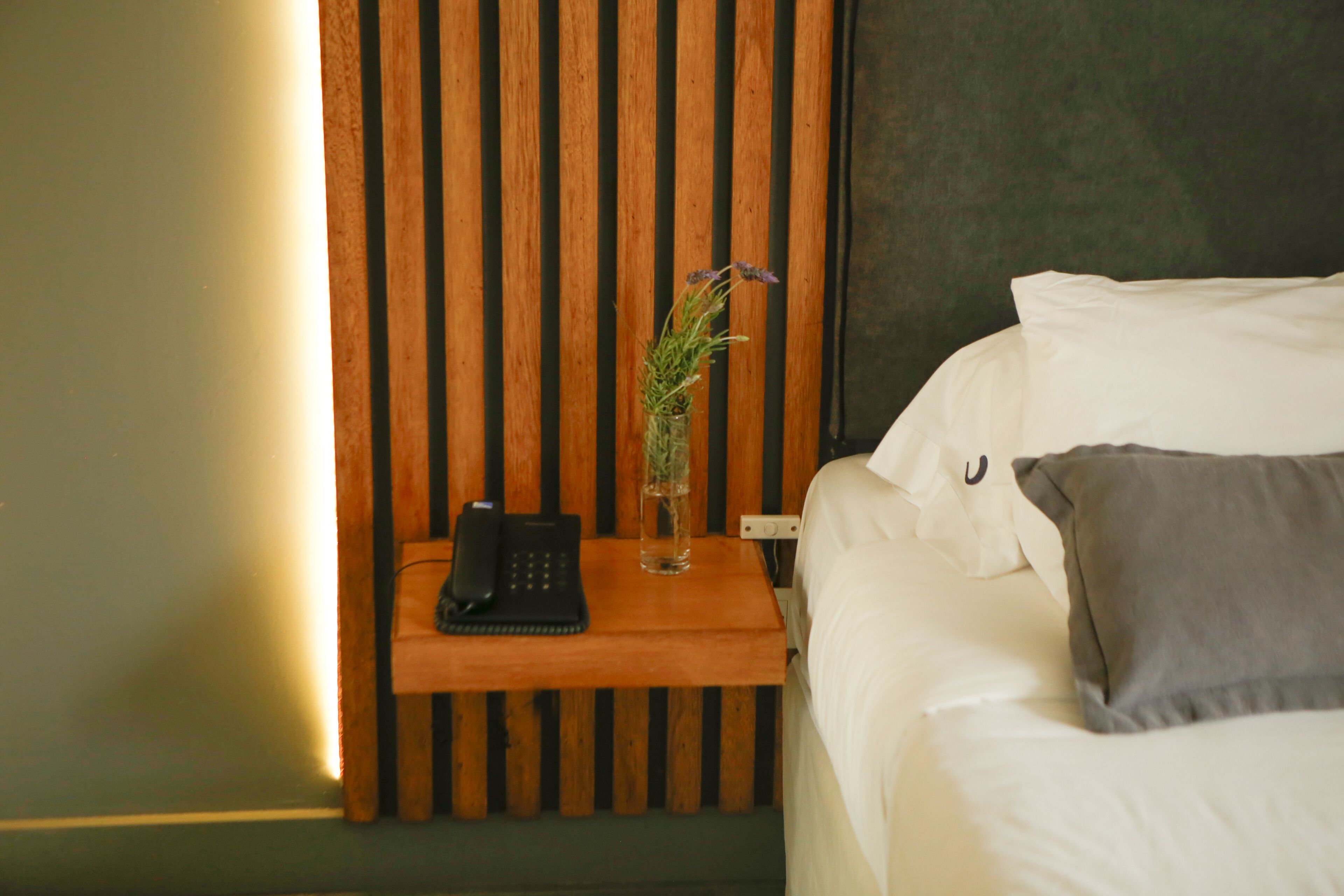 A neatly made bed with white linens and a gray pillow, a bedside table with a phone and a vase of flowers, and a wooden slat wall behind the table.