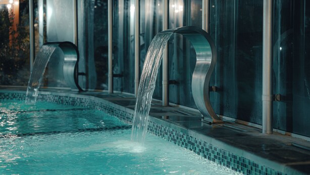 A pool with water cascading from a curved structure into the water. - Destinar Apart Hotel (Carilo)