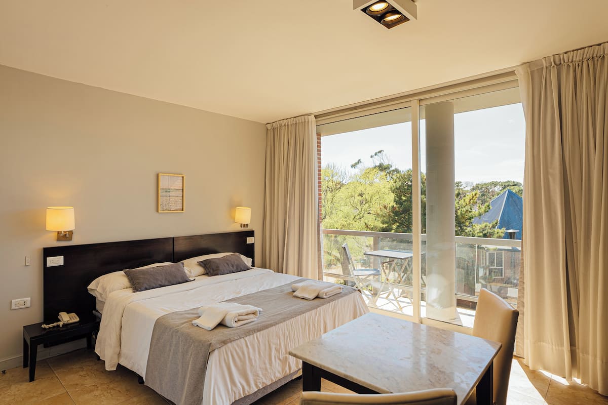 A hotel room with a bed, bedside lamps, a table, and a balcony with a view of trees and a building.