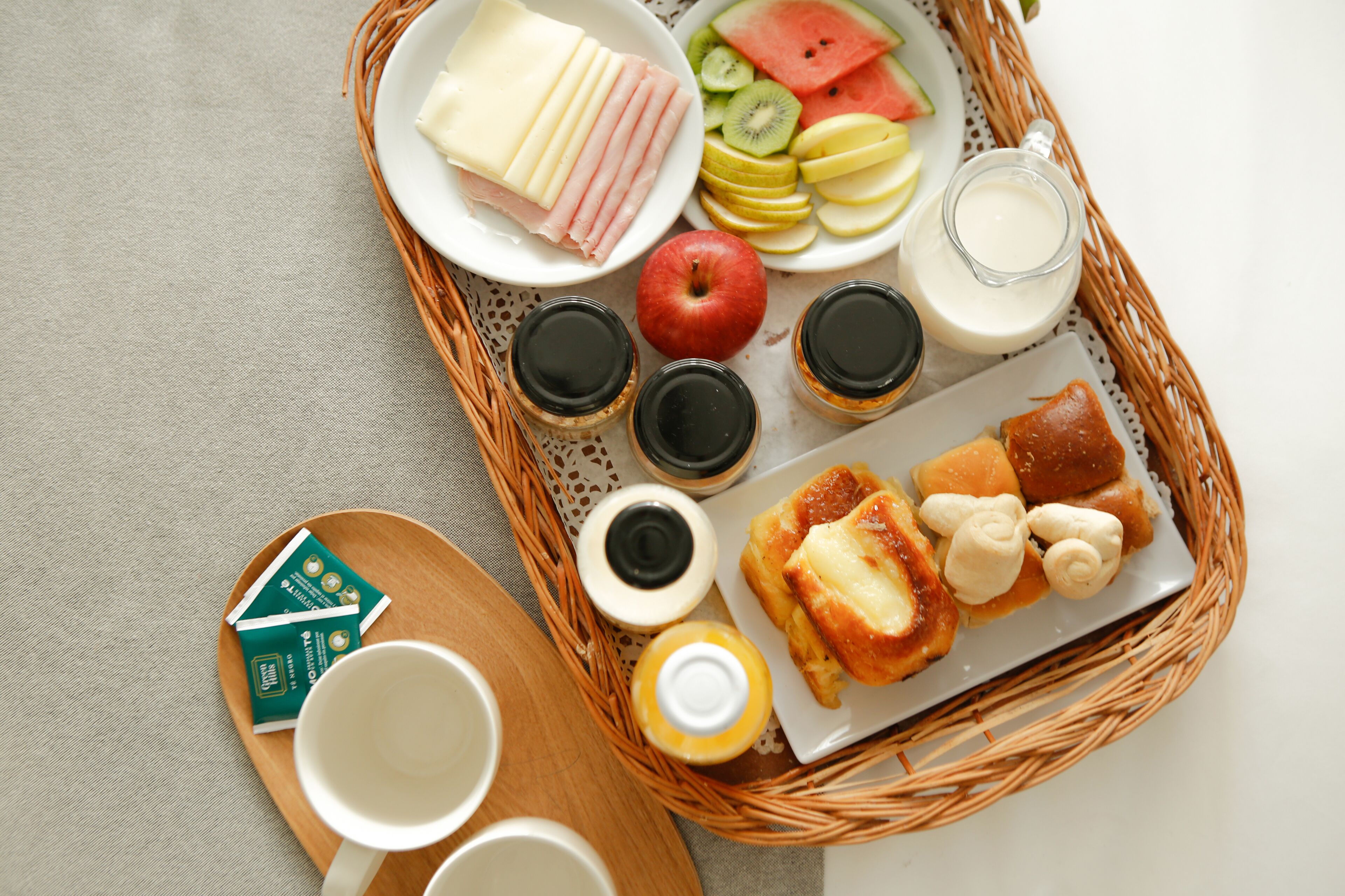 Bandeja de desayuno con jamón, queso, frutas, yogur y pastelería sobre un tapete tejido.