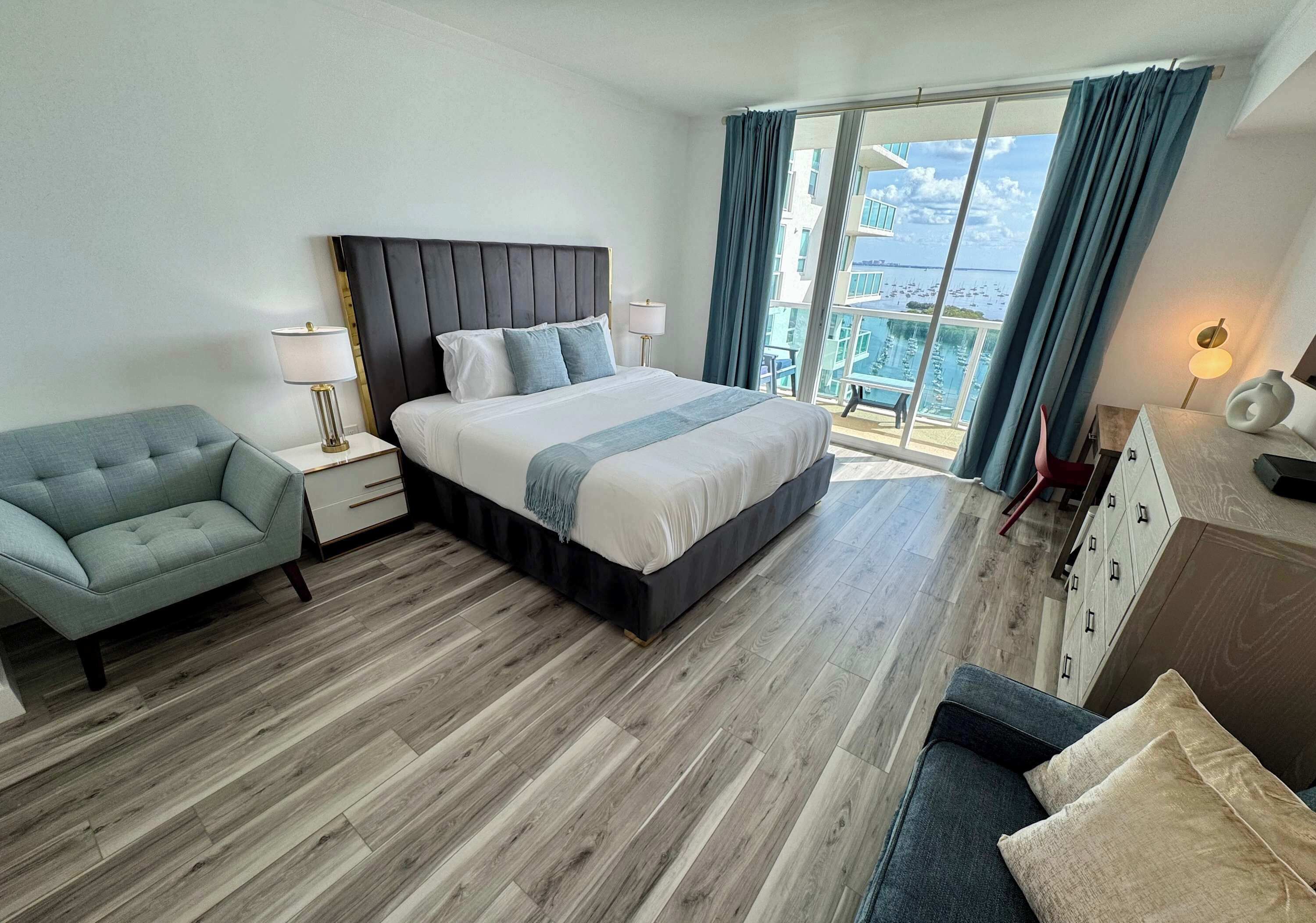 Habitación de hotel con una cama grande, un sofá, una mesita de noche, una lámpara, un aparador y un balcón con vistas al mar.