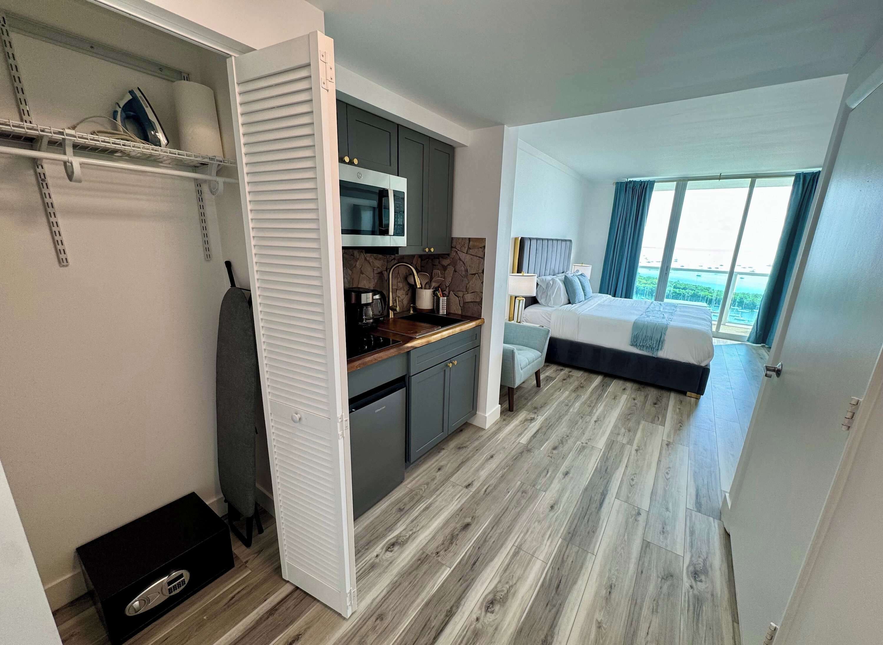 A modern hotel room with a kitchen area, a bed with a view, and a sliding door closet.