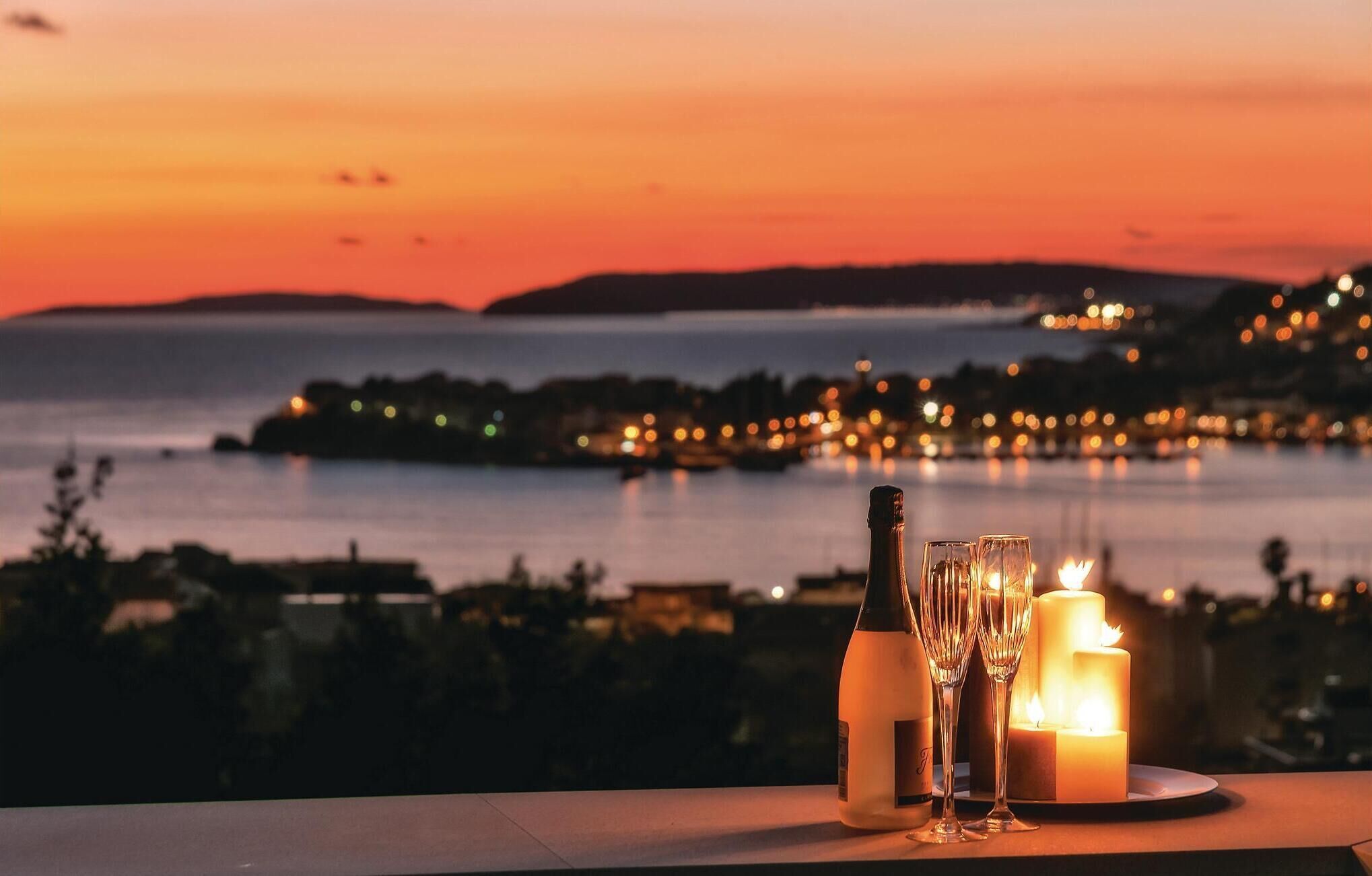 Une table avec une bouteille, deux verres et une bougie allumée, donnant sur une vue côtière au coucher du soleil.