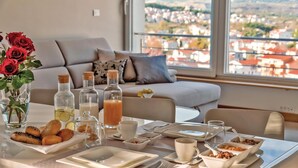 A breakfast setting with a table set for two, including plates, cutlery, and a variety of food items. There are also several bottles, possibly containing beverages or condiments. A vase with red roses is visible on the table, and the room has a view of a cityscape through large windows.