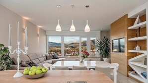 A modern living room with a sofa, dining table, and a view of a mountainous landscape through large windows.