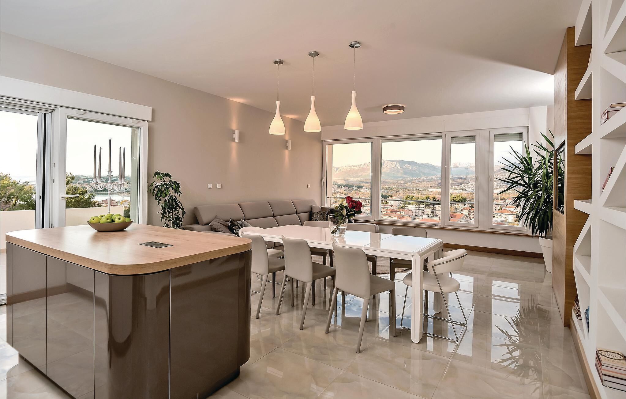 A modern kitchen with a central island, dining table, and chairs. There are pendant lights, a sofa, and large windows with a view of the city.