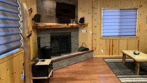 Una habitación acogedora con chimenea de piedra, paredes de madera, televisor de pantalla plana y una ventana con persianas.