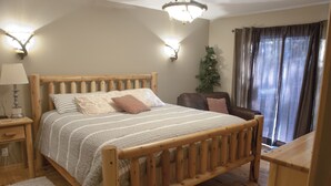 Una cama de madera con cabecera, una mesita de noche con lámpara, una silla, una pequeña mesa y un árbol de Navidad.