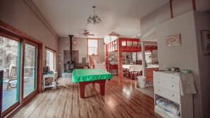 Una habitación amplia con mesa de billar, piso de madera, ventilador de techo y vistas al exterior a través de grandes ventanales.