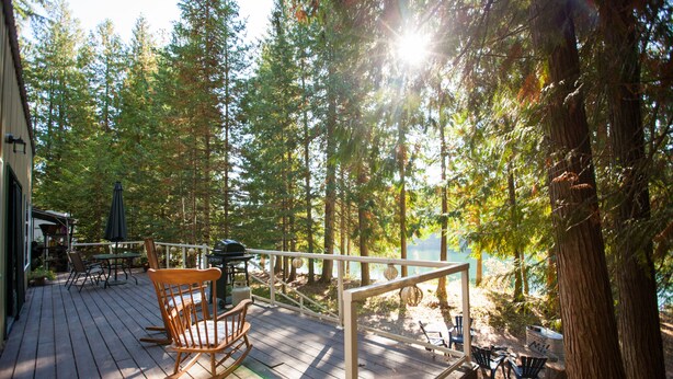 Una terraza de madera con mobiliario exterior, con vista a un bosque y una masa de agua.