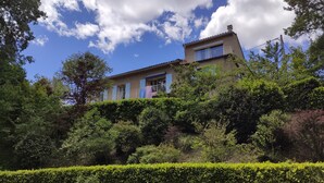 Un edificio de dos pisos con techo plano, rodeado de abundante vegetación y un cielo despejado.