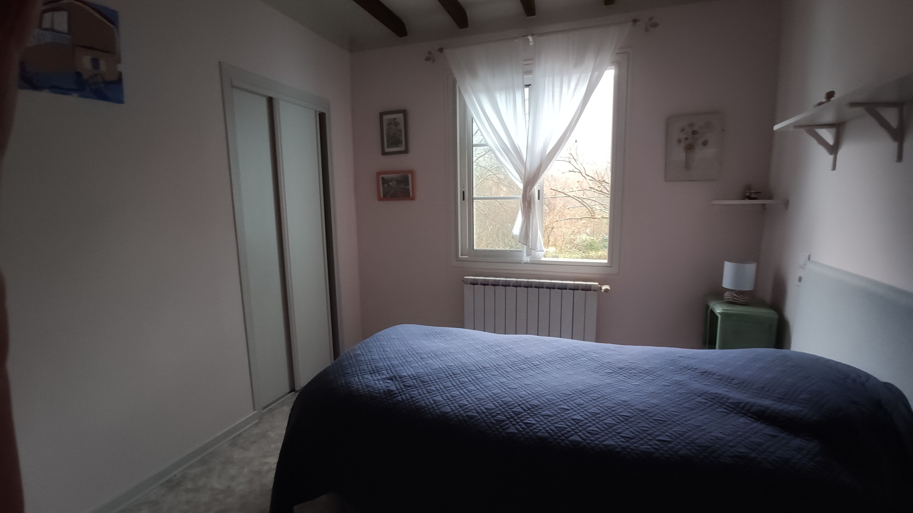 Un dormitorio con cama, ventana con cortinas, radiador y una pequeña mesa con lámpara.