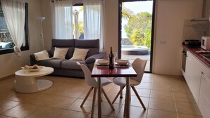 Ein modernes Wohnzimmer mit einem grauen Sofa, einem Esstisch mit Stühlen, einem kleinen runden Tisch mit einer Schüssel Snacks und einer sichtbaren Whirlwanne durch die Glastür.