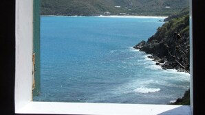 Vista costeira através de uma janela arqueada, com mar azul cristalino, costa rochosa e colinas verdes ao fundo.