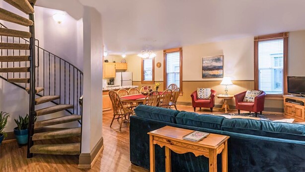 A living room with a spiral staircase, a dining area with wooden chairs, a blue sofa, and a TV. - Oak Sherwin House Near OSF (Ashland)