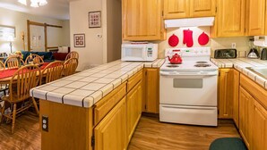 A compact kitchen with wooden cabinets, a white stove, a microwave, and a dining area with a red tablecloth. - Oak Sherwin House Near OSF (Ashland)