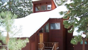 A wooden cabin with a steep staircase leading to a door, surrounded by snow and trees.