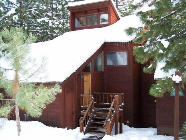 A wooden cabin with a steep staircase leading to a door, surrounded by snow and trees.