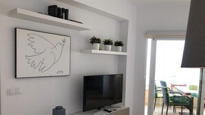 Un soggiorno moderno con una TV a schermo piatto su un mobile bianco, un quadro incorniciato sopra la TV e una piccola pianta in vaso su uno scaffale.
