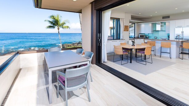 A modern kitchen with white cabinets and a large island, a dining area with wicker chairs, and a view of the ocean. - Beachfront  / Corner  / Renovated / 2nd floor condo unit: F-204 (Wailea-Makena)
