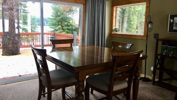 A dining area with a wooden table and chairs, large windows with curtains, a view of trees and a deck outside, and a lamp on the wall.