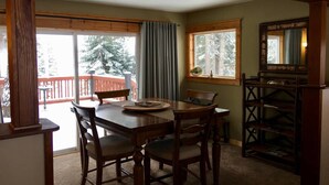 A dining area with a wooden table and chairs, a window with a view of a snowy deck, and a wooden bookshelf.