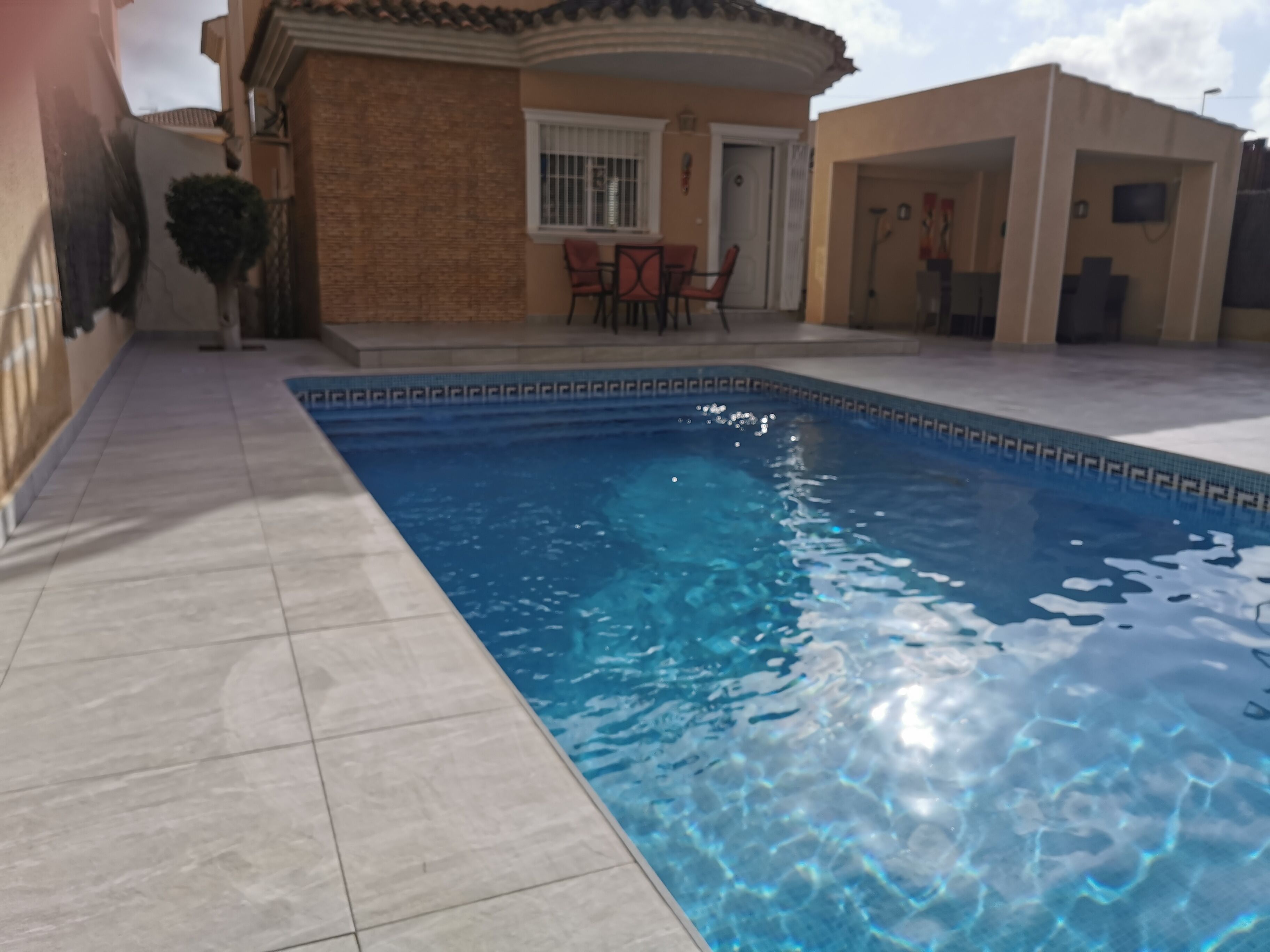 Una piscina con bordi piastrellati, un'area patio con sedie e un piccolo albero.