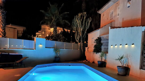 Una piscina al aire libre con un diseño geométrico de azulejos, rodeada de plantas en macetas y una pared blanca con barandilla decorativa.