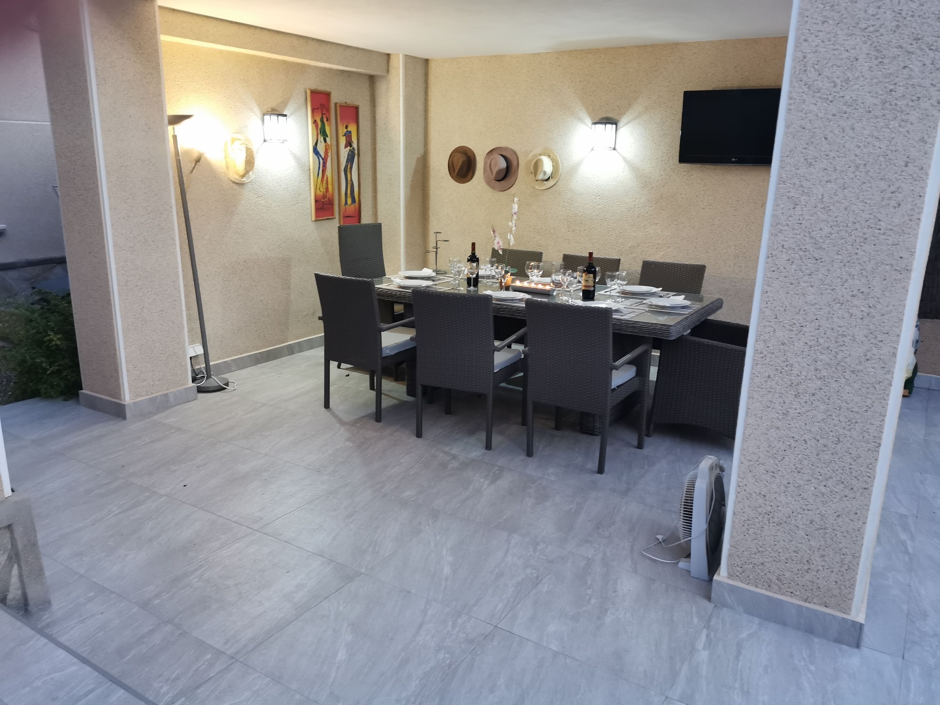 Un coin repas avec une table dressée pour quatre personnes, entourée de chaises en osier, d’un lampadaire et de luminaires fixés au mur.
