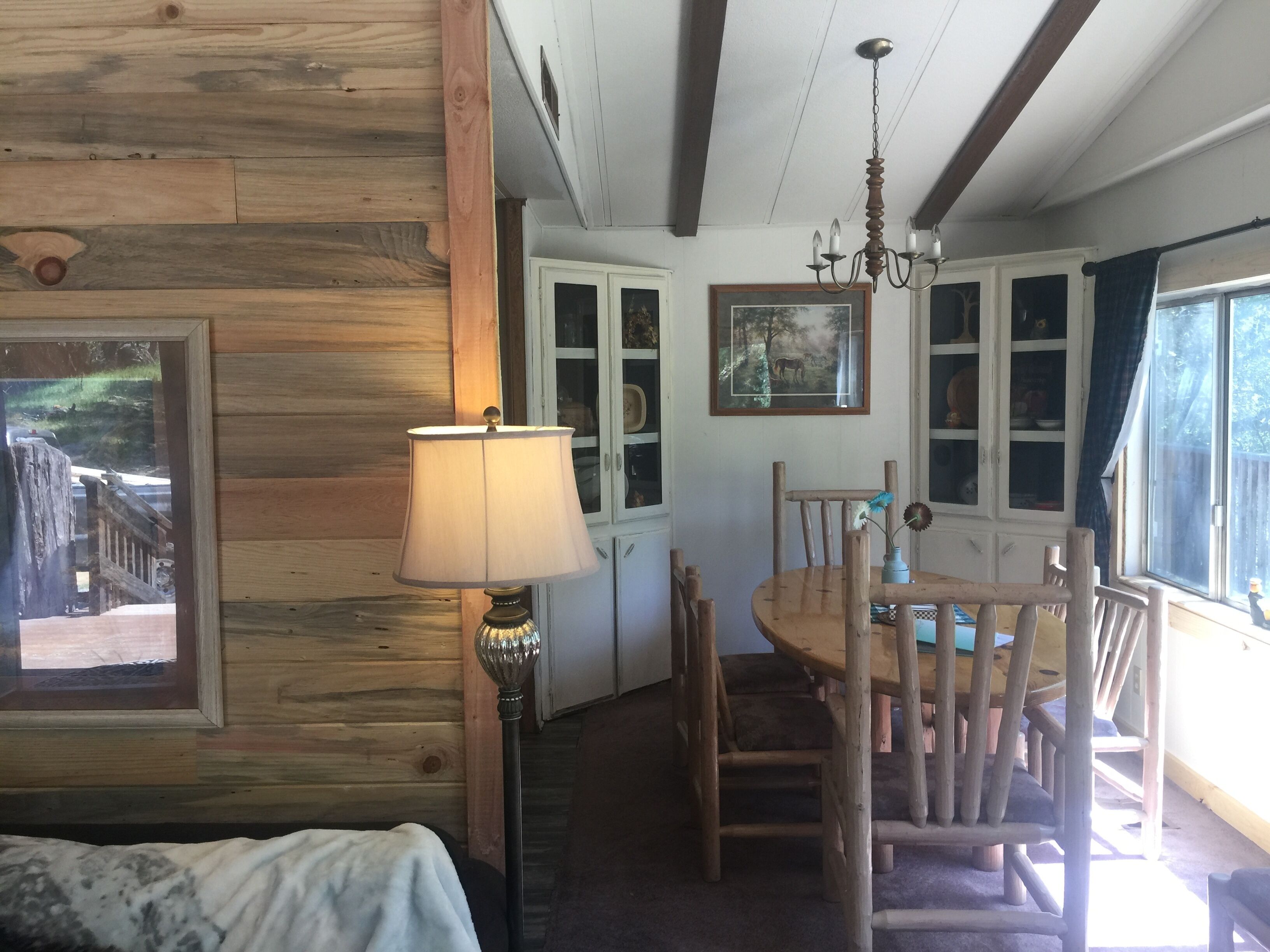 A rustic cabin interior with a wooden dining table, chairs, a chandelier, and a framed picture on the wall.