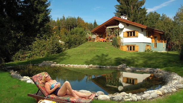 A woman relaxing on a lounge chair by a pond, with a house in the background. - Quietly located, luxurious 4-stars-country house with private pool (Schiefling am See)
