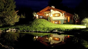 A two-story house with a sloped roof, a satellite dish, and a pond in front. - Quietly located, luxurious 4-stars-country house with private pool (Schiefling am See)