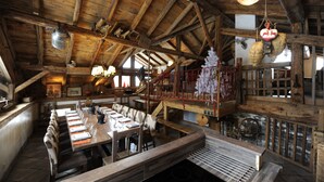 Un intérieur rustique en bois, avec une longue table à manger dressée pour un repas, un escalier et un luminaire suspendu.