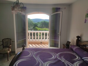 Relax in a spacious room with a large bed, wooden chair, and balcony offering views of lush greenery and hills.