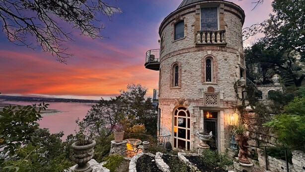 A historic stone tower with a balcony, surrounded by lush greenery and a view of a body of water during sunset.