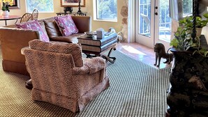 A cozy living room with a leather sofa, patterned armchair, and a coffee table. There are framed artworks on the walls and a large window with a view of greenery.