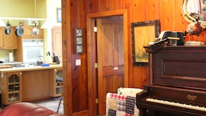 A wooden-paneled room with a piano, a mirror, and a kitchen area visible through an open door. - Grandma's House: Riverfront, Gorgeous View, Beach/Swimming/Canoeing Dog Friendly (Monte Rio)