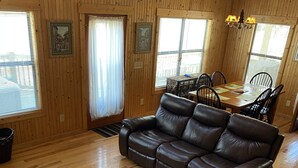 A cozy cabin interior with wooden walls, a leather sofa, a dining table with chairs, and a staircase with a wooden railing.