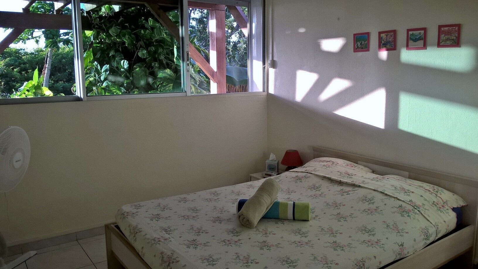 A bedroom with a bed, a fan, a window, and a wall with framed pictures.